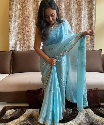 A woman showcasing a blue saree with embroidery and a scalloped border, standing in a room with a sofa and curtains in the background.