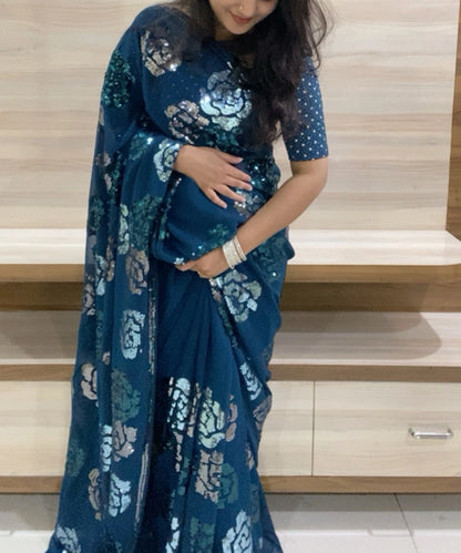 A woman wearing a blue floral saree with a matching blouse, holding pleats of the saree.
