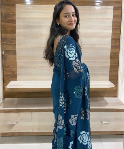 Woman wearing a blue saree with white patterns in a room with wooden furniture.